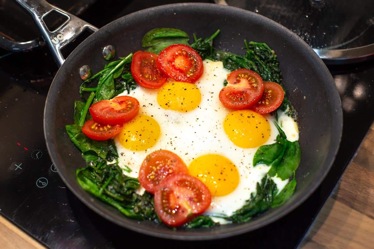 Eggs frying in a pan with sliced tomatoes and spinach.