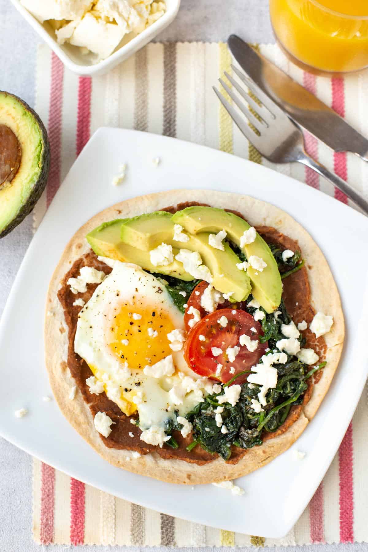 A breakfast tostada on a plate, topped with sliced avocado and crumbled feta.