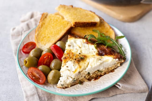 A slice of ricotta frittata on a plate served with buttered toast, olives and tomatoes.