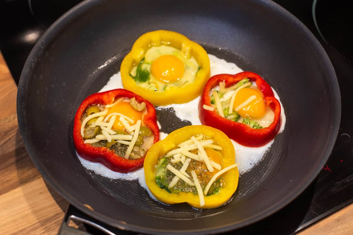Uncooked eggs inside pepper slices in a frying pan, topped with grated cheese.