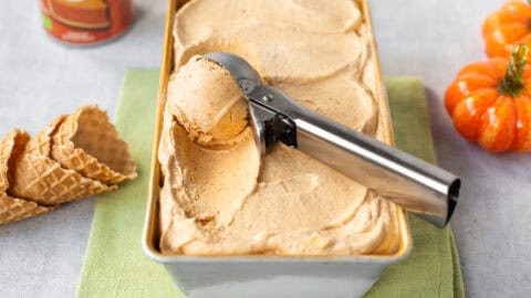 A metal ice cream scoop taking a spoonful of pumpkin pie ice cream from a loaf tin.