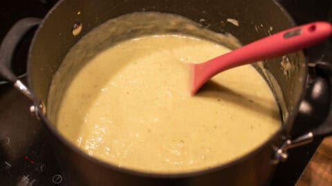 Creamy cauliflower cheese soup in a large saucepan with a pink spoon.