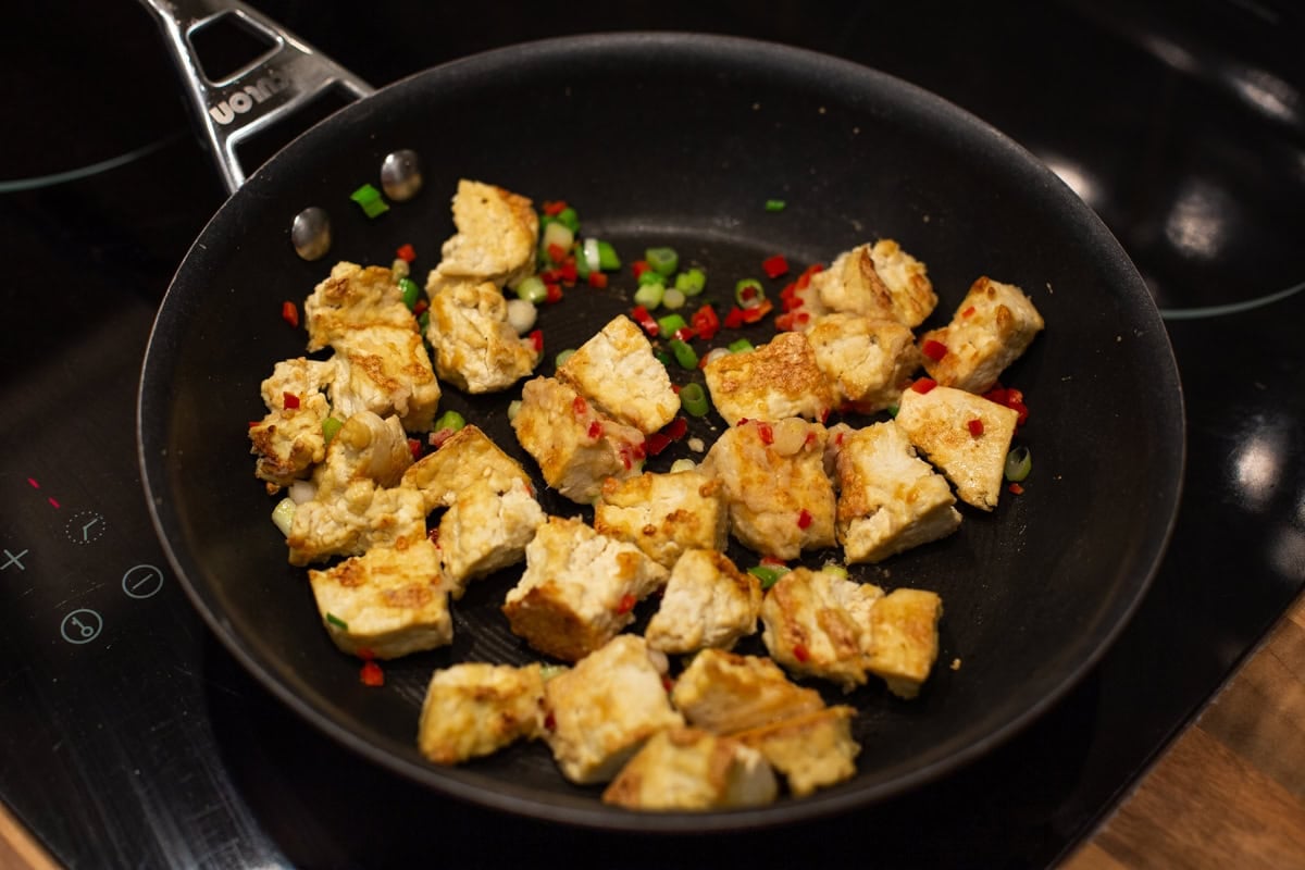 Crispy tofu cooking in a frying pan with chilli and spring onions.