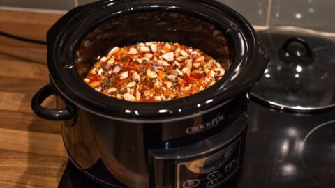 A slow cooker filled with uncooked ingredients for lentil bolognese, including lentils and mushrooms.