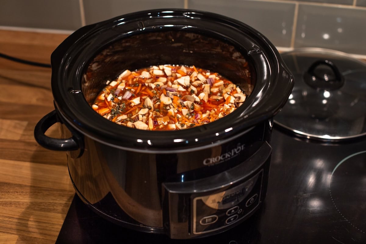 A slow cooker filled with uncooked ingredients for lentil bolognese, including lentils and mushrooms.