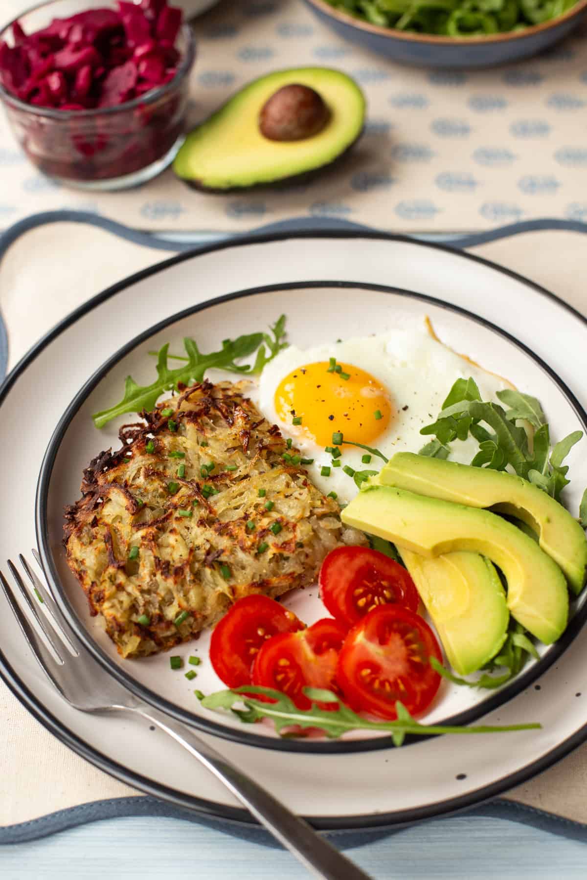 A portion of air fryer hash browns on a plate with a fried egg, tomatoes and avocado.