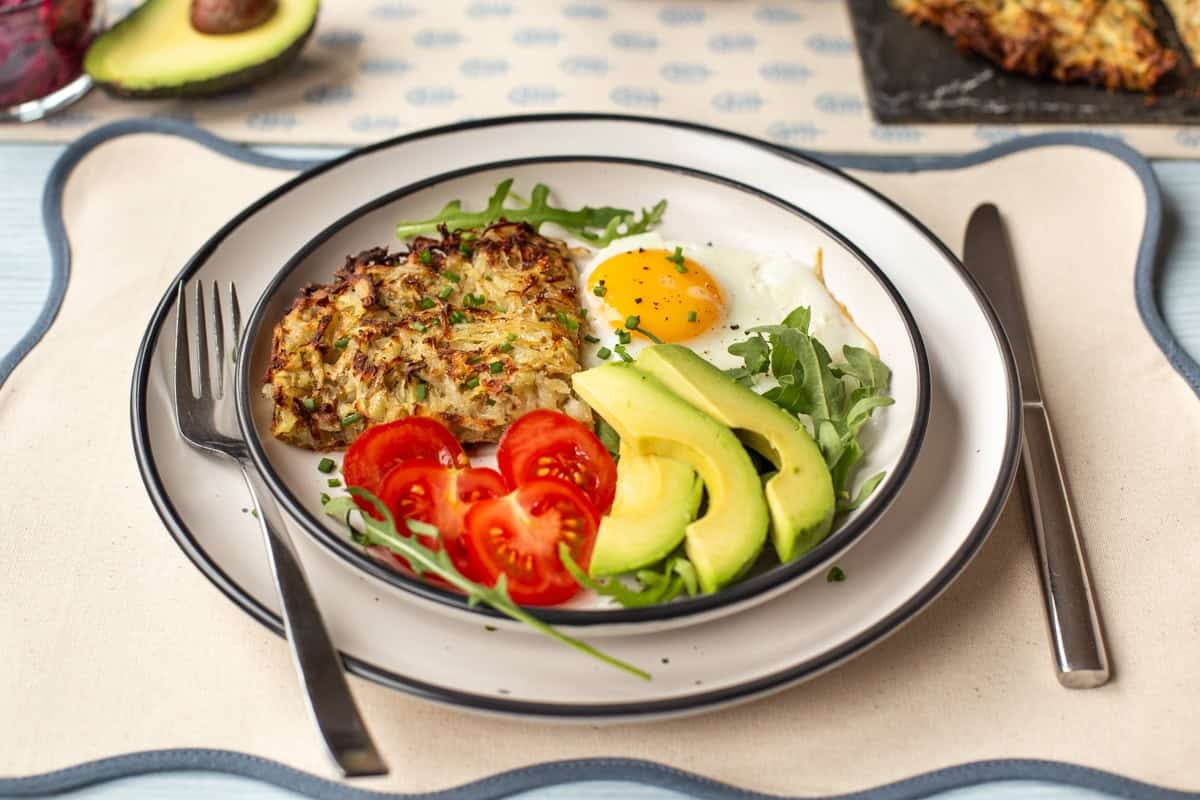 A portion of air fryer hash browns on a plate with a fried egg, tomatoes and avocado.