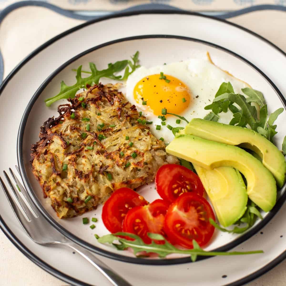 A portion of air fryer hash browns on a plate with a fried egg, tomatoes and avocado.