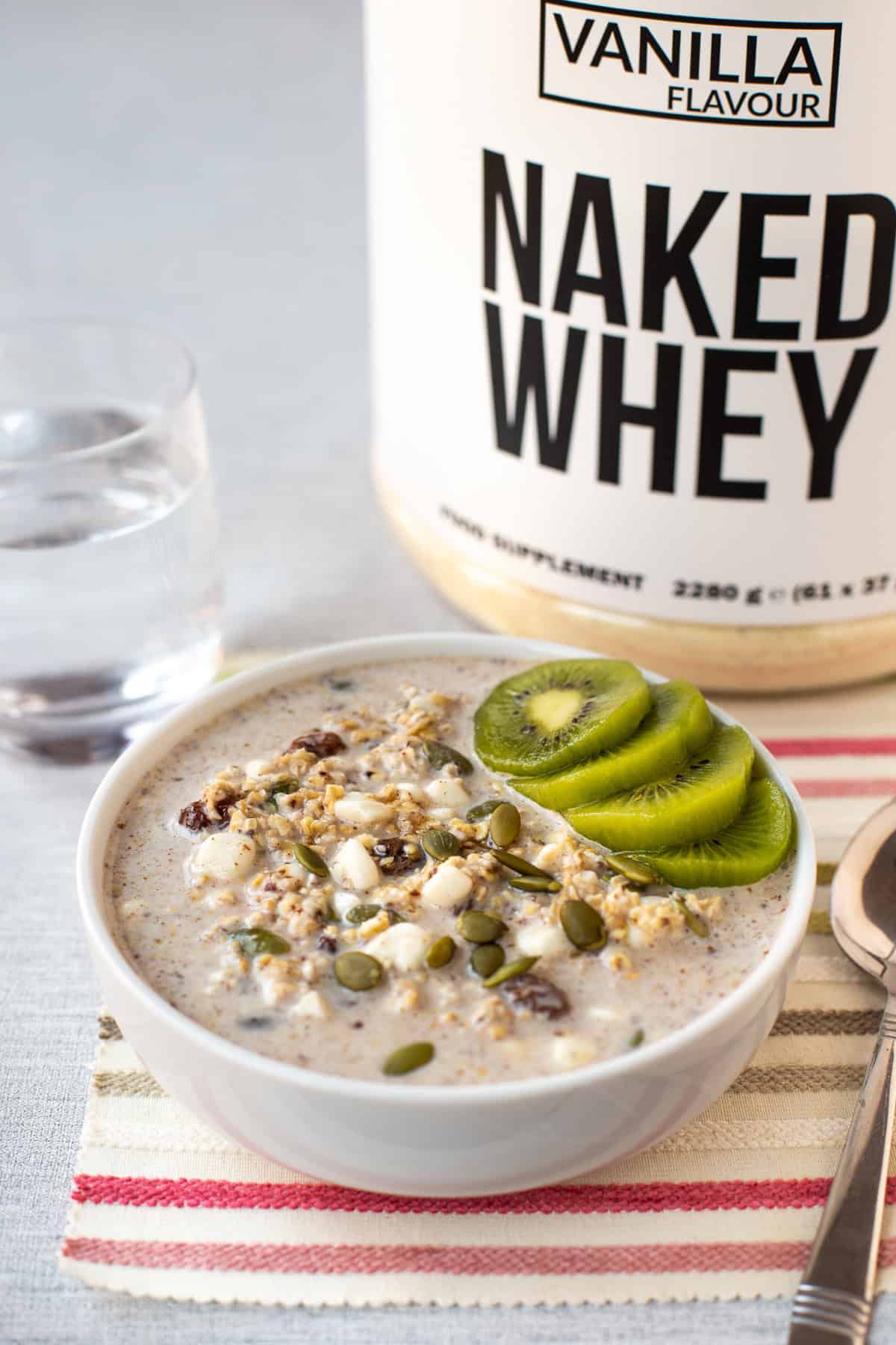 A bowl of high protein overnight oats in front of a large tub of vanilla flavour whey protein powder.