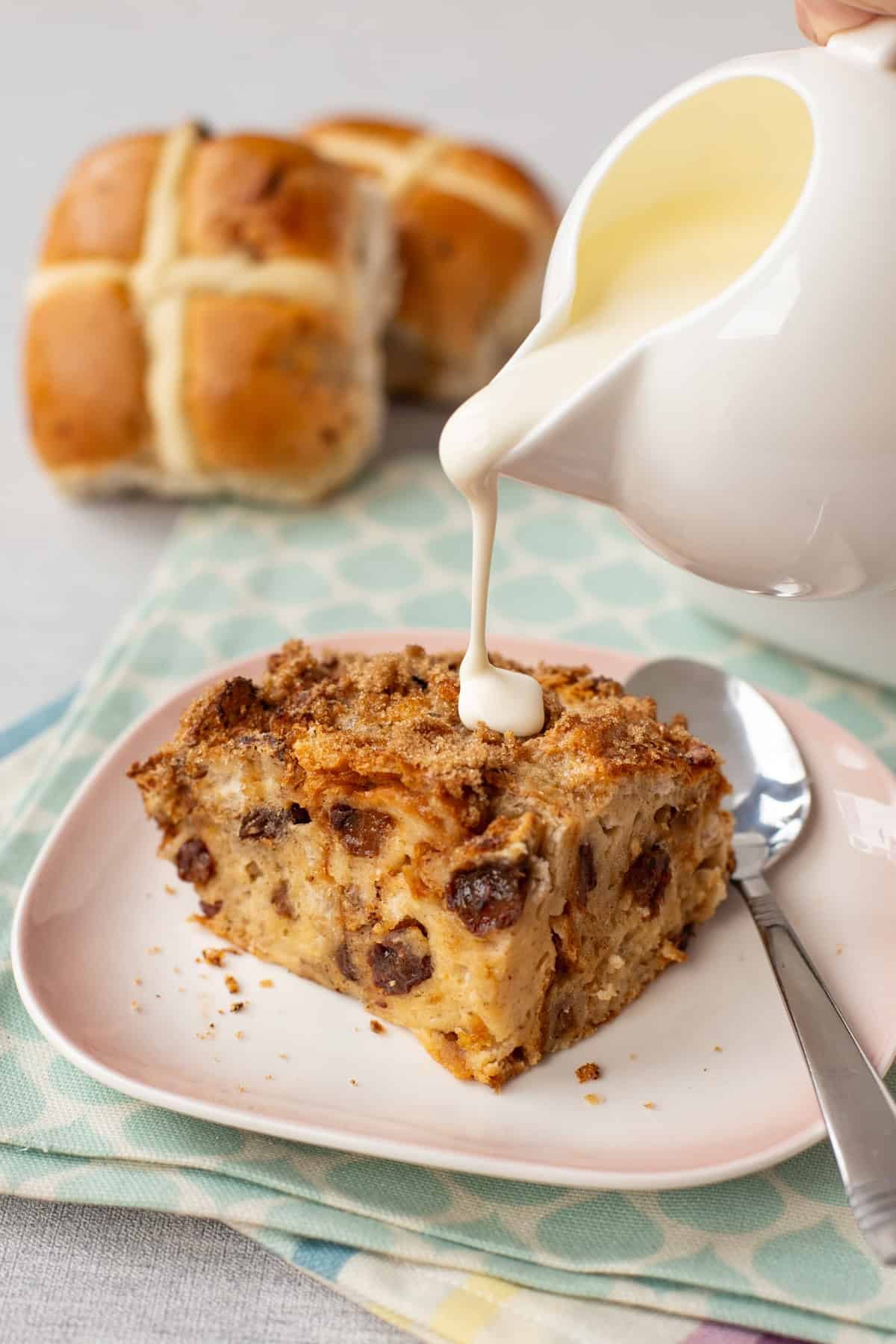 Cream being poured over a piece of hot cross bun bread and butter pudding on a plate.