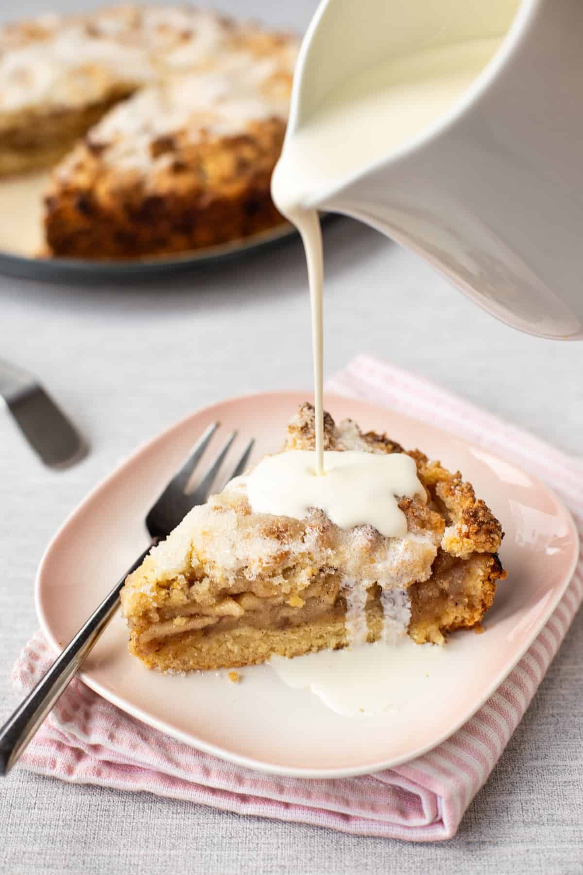 A slice of easy apple crumble cake on a plate with cream being poured on top.