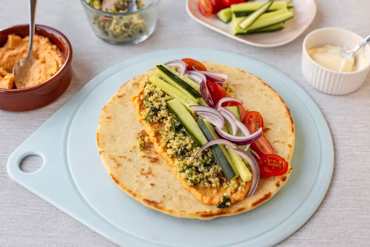 A flatbread laid on a board topped with hummus, tabbouleh and vegetables.