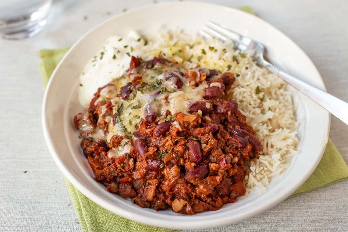 A portion of kidney bean chilli in a bowl with rice, cheese and sour cream.