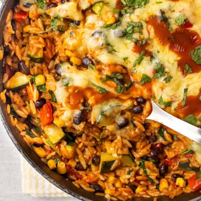 A large spoon taking a scoop from a pan of cheesy enchilada orzo.