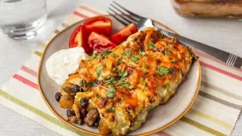 A portion of cheesy potato enchiladas on a plate with sour cream and tomatoes.