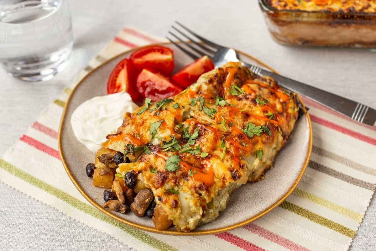 A portion of cheesy potato enchiladas on a plate with sour cream and tomatoes.