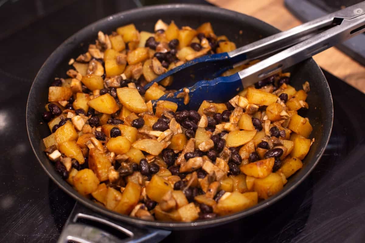 Potato, black bean and mushroom mixture cooking in a frying pan.