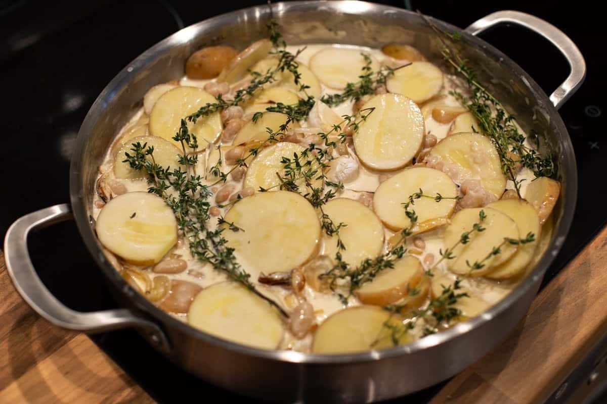 Sliced potatoes, beans and cream with thyme and garlic in a metal pan.